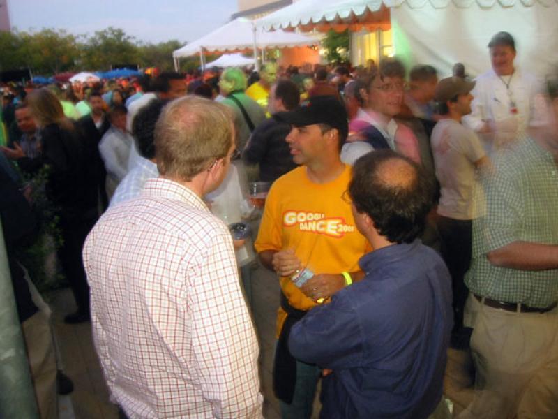 A large crowd of mostly young men mingle in and around marquis tents set up for the Google Dance in 2005. In the center, a man wearing a baseball cap and a t-shirt with the event logo talks to 3 other men. 